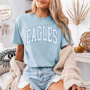 May include: A light blue short-sleeve t-shirt featuring the word EAGLES in a white and blue striped collegiate font across the chest. The shirt is made of 100% cotton and is available in more than 10 colors. The text Chambray is visible.