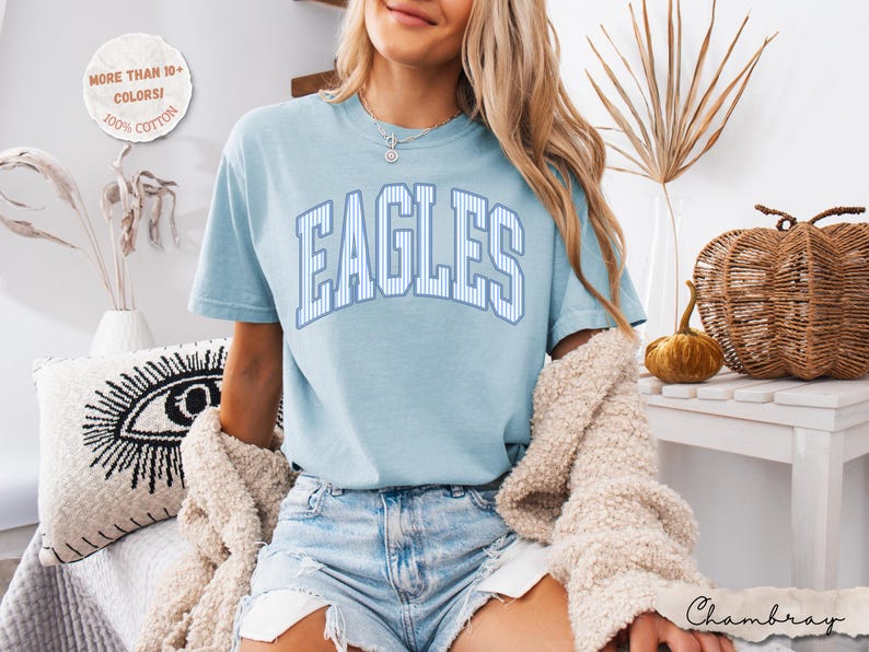 May include: A light blue short-sleeve t-shirt featuring the word EAGLES in a white and blue striped collegiate font across the chest. The shirt is made of 100% cotton and is available in more than 10 colors. The text Chambray is visible.