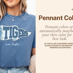 May include: A blue t-shirt featuring a white and blue checkered pennant graphic with the text TIGERS and the name mrs. taylor written below. The right side contains text about Pennant Color and personalization instructions in brown font.
