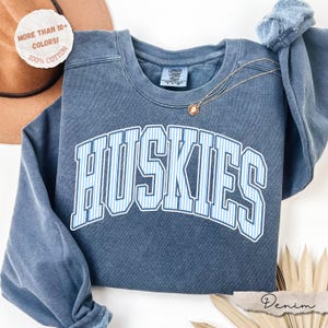 May include: A blue crewneck sweatshirt featuring the word HUSKIES in a white and light blue striped font across the chest. The garment has a faded, vintage-style wash and a Comfort Colors label at the inner neckline. A thin gold chain necklace rests on the fabric.