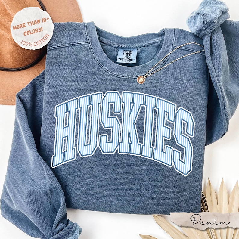 May include: A blue crewneck sweatshirt featuring the word HUSKIES in a white and light blue striped font across the chest. The garment has a faded, vintage-style wash and a Comfort Colors label at the inner neckline. A thin gold chain necklace rests on the fabric.