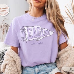 May include: A light purple short-sleeve t-shirt featuring a white and light purple checkered pennant graphic with the text "EAGLES" in large purple letters and "mrs. taylor" in smaller script below. The shirt has a crew neckline and a relaxed fit.