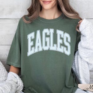 May include: A moss green short-sleeve t-shirt featuring the word EAGLES printed in a large, arched, white-outlined font with a light blue and white vertical pinstripe pattern inside the letters, centered on the chest of the garment.