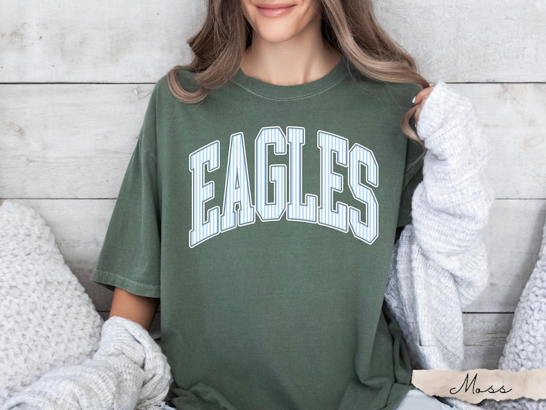 May include: A moss green short-sleeve t-shirt featuring the word EAGLES printed in a large, arched, white-outlined font with a light blue and white vertical pinstripe pattern inside the letters, centered on the chest of the garment.