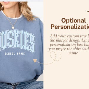 May include: A blue crewneck sweatshirt featuring the word HUSKIES in a white and blue striped collegiate font with the text SCHOOL NAME underneath. The right side displays the text Optional Personalization! Add your custom text below the mascot design! Leave the personalization box blank if you prefer the shirt without a name.