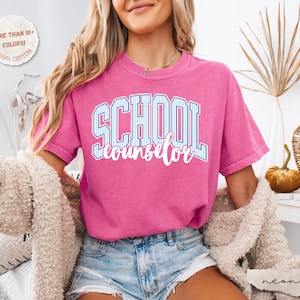 Puede incluir: Una camiseta de manga corta de color rosa ne&oacute;n con la palabra SCHOOL en una fuente universitaria de rayas azules y blancas, con la palabra counselor escrita debajo en una fuente cursiva blanca. La camiseta tiene cuello redondo y corte holgado.