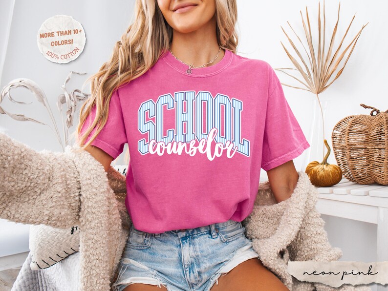 Puede incluir: Una camiseta de manga corta de color rosa ne&oacute;n con la palabra SCHOOL en una fuente universitaria de rayas azules y blancas, con la palabra counselor escrita debajo en una fuente cursiva blanca. La camiseta tiene cuello redondo y corte holgado.