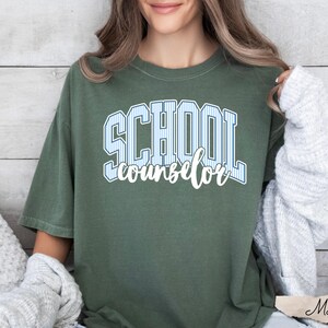 Puede incluir: Una camiseta de manga corta color verde oliva con el texto SCHOOL en una fuente universitaria de rayas azul claro y blanco, con la palabra counselor escrita debajo en letra cursiva blanca. La camiseta tiene cuello redondo y corte holgado.