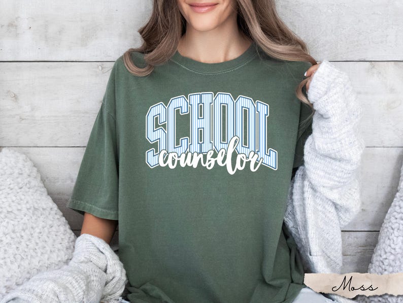Puede incluir: Una camiseta de manga corta color verde oliva con el texto SCHOOL en una fuente universitaria de rayas azul claro y blanco, con la palabra counselor escrita debajo en letra cursiva blanca. La camiseta tiene cuello redondo y corte holgado.