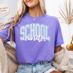 Puede incluir: Una camiseta violeta de manga corta con el texto "SCHOOL counselor" impreso en la parte delantera. La palabra "SCHOOL" aparece en una fuente de bloque grande con rayas azul claro y blancas, y "counselor" debajo en letra cursiva blanca.