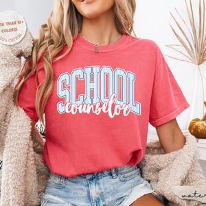 Puede incluir: Una camiseta de manga corta color coral con el texto SCHOOL en rayas verticales azules y blancas y la palabra counselor en letra cursiva blanca. Una insignia circular en la esquina superior izquierda indica MORE THAN 10+ COLORS! 100% COTTON.