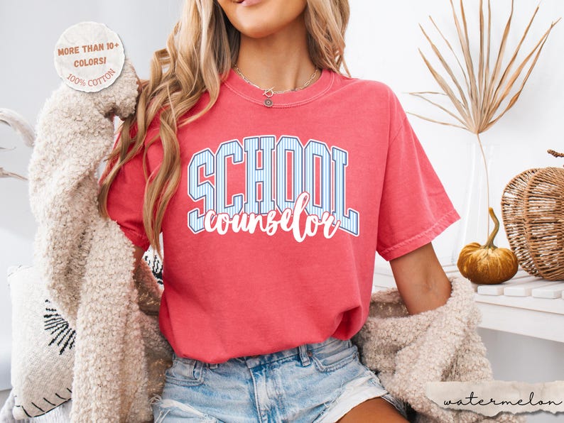 Puede incluir: Una camiseta de manga corta color coral con el texto SCHOOL en rayas verticales azules y blancas y la palabra counselor en letra cursiva blanca. Una insignia circular en la esquina superior izquierda indica MORE THAN 10+ COLORS! 100% COTTON.
