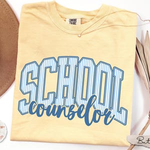 Puede incluir: Una camiseta de manga corta de color amarillo p&aacute;lido con el texto SCHOOL counselor en una fuente universitaria de rayas azules y blancas. La camiseta tiene una etiqueta de la marca Comfort Colors en el cuello y se muestra con un sombrero marr&oacute;n.