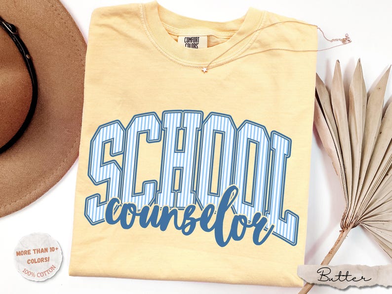 Puede incluir: Una camiseta de manga corta de color amarillo p&aacute;lido con el texto SCHOOL counselor en una fuente universitaria de rayas azules y blancas. La camiseta tiene una etiqueta de la marca Comfort Colors en el cuello y se muestra con un sombrero marr&oacute;n.