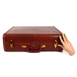 May include: A vintage brown leather suitcase with gold-toned hardware, including latches and a handle. The suitcase has a rectangular shape with rounded corners and a keyhole lock. The stitching is visible along the edges.