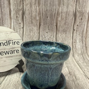 May include: A handcrafted ceramic flower pot with a matching saucer. The pot is glazed in shades of blue and teal, with a glossy finish. The pot has a classic shape with a slightly flared rim. The saucer is round and fits snugly under the pot.