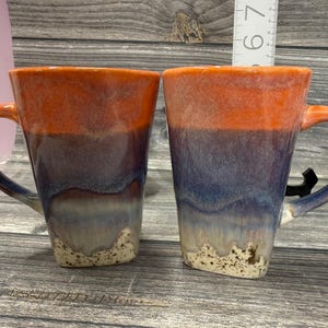 May include: Two ceramic mugs with a square base and a curved handle. The mugs have a gradient design, transitioning from orange at the top to blue and white at the bottom. The handles are orange. The mugs are on a wooden surface.