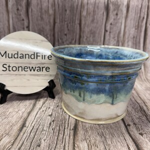 May include: A handcrafted stoneware bowl with a blue and white glaze. The bowl features a textured exterior and a smooth interior. The text "MudandFire Stoneware" is visible on a white circular sign in the background.