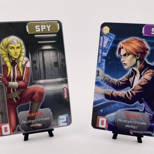 May include: Two trading cards featuring illustrations of characters. One card shows a woman in a red and white outfit, the other a person in a brown jacket. Both cards have the word "SPY" and other text. The cards are displayed on small black stands.
