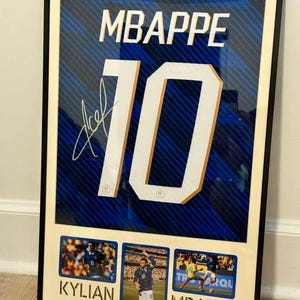 May include: Framed artwork featuring a blue and white soccer jersey with the name "MBAPPE" and the number "10" in large white letters. Includes a signature and three small action photos of the player. The frame is black.