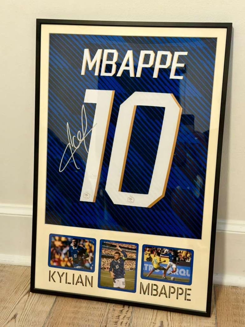 May include: Framed artwork featuring a blue and white soccer jersey with the name "MBAPPE" and the number "10" in large white letters. Includes a signature and three small action photos of the player. The frame is black.