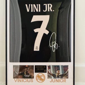Puede incluir: Camiseta de fútbol en blanco y negro enmarcada con el nombre "VINI JR." y el número "7" en blanco. Incluye una firma e imágenes del jugador. La parte inferior del marco tiene el nombre "VINICIUS JUNIOR" en dorado.