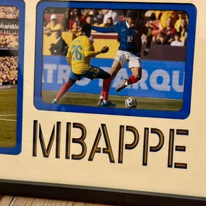 May include: A framed photo featuring a soccer match with players in blue and yellow uniforms. The word "MBAPPE" is printed in large letters below the photo. The frame has a blue border and a light beige background.