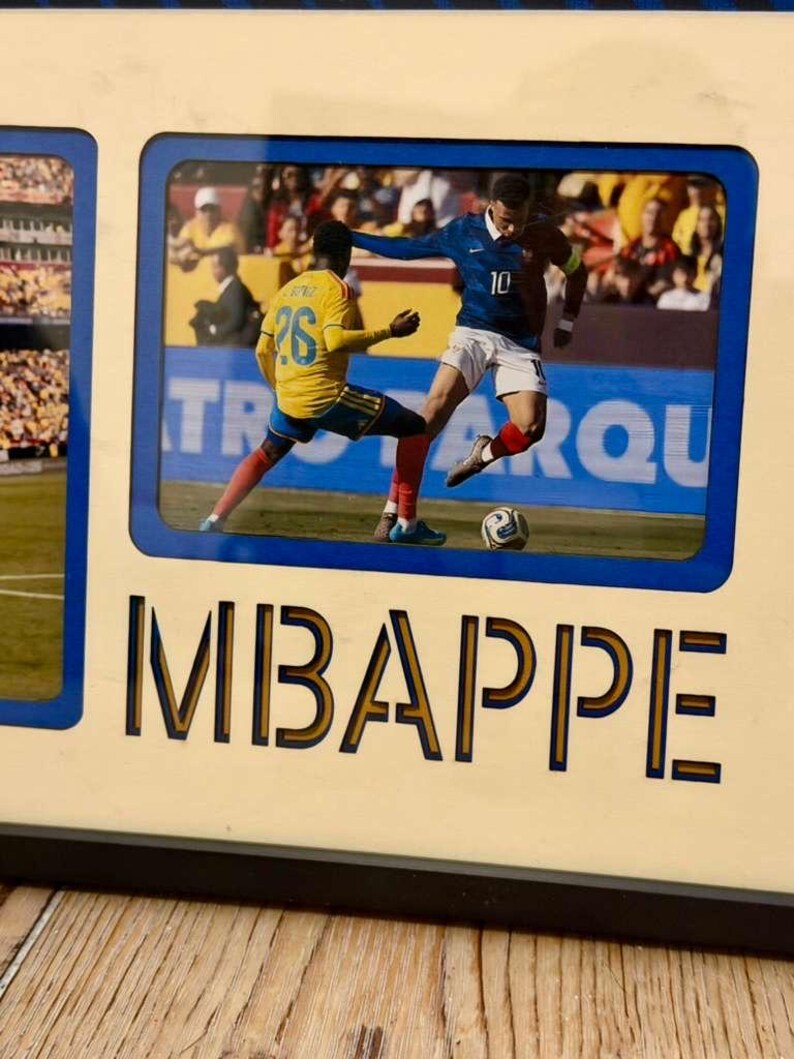 May include: A framed photo featuring a soccer match with players in blue and yellow uniforms. The word "MBAPPE" is printed in large letters below the photo. The frame has a blue border and a light beige background.