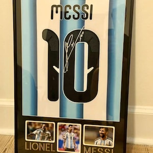 May include: Framed artwork featuring a soccer jersey with the name "MESSI" and the number "10". The jersey has blue and white stripes. Below are three small photos of Lionel Messi. The frame is black.