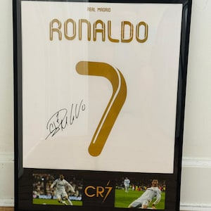 May include: Framed print featuring a white jersey with "RONALDO" and the number "7" in gold. Includes a signature and images of the player. The words "REAL MADRID" and "CR7" are also visible.