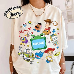 May include: Ivory-colored t-shirt featuring colorful cartoon characters. The shirt includes the text "COMFORT COLORS" and "Ivory" in a circular design. Various cartoon characters are printed on the shirt, along with the word "NAME" in a green frog-shaped frame.