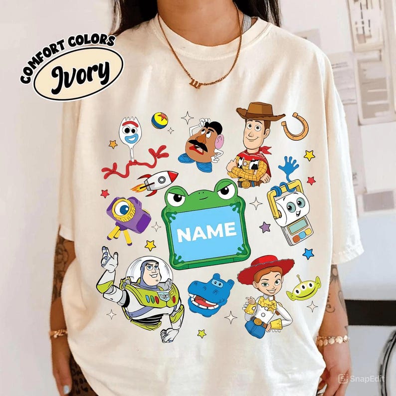 May include: Ivory-colored t-shirt featuring colorful cartoon characters. The shirt includes the text "COMFORT COLORS" and "Ivory" in a circular design. Various cartoon characters are printed on the shirt, along with the word "NAME" in a green frog-shaped frame.