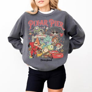May include: Dark gray sweatshirt featuring a colorful graphic of Pixar characters and the words "Pixar Pier" and "Disneyland" in red and white. The design includes characters from Toy Story, Monsters, Inc., and Cars.