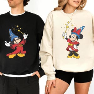 May include: Black and cream sweatshirts featuring Mickey and Minnie Mouse as wizards. Mickey wears a red robe and blue wizard hat, Minnie a red dress and hat. Both hold wands with star accents, perfect for Disney fans.