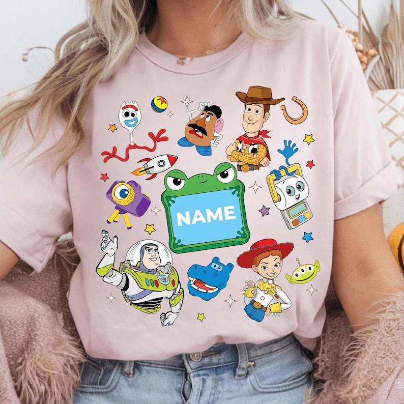 May include: A light pink t-shirt featuring a colorful graphic of cartoon characters. The design includes a green frog with the word "NAME" inside, a cowboy, a space ranger, and other characters. The shirt is a crew neck.