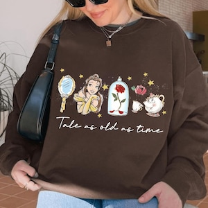 Könnte beinhalten: Ein braunes Sweatshirt mit einem Design aus dem Film Die Schöne und das Biest. Das Design zeigt Belle, eine Rose in einem Glaskasten und den Text "Tale as old as time".