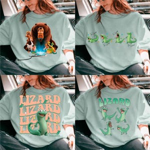 May include: Four light blue sweatshirts with different graphic designs. One features a bear and other cartoon characters. Another has green lizards with the text "The Legend". The other two have the word "Lizard" with a cartoon lizard.