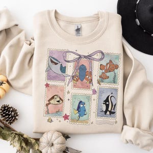 May include: Beige crewneck sweatshirt featuring a colorful graphic of characters from the animated film "Finding Nemo" in postage stamp designs. The design includes a shark, clownfish, and other sea creatures. A purple bow is at the top of the design.