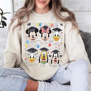 May include: Cream-colored sweatshirt featuring a colorful graphic of Disney characters Mickey Mouse, Minnie Mouse, Donald Duck, Daisy Duck, Pluto, and Goofy, all wearing graduation caps. The design includes balloons, stars, and the characters' names.