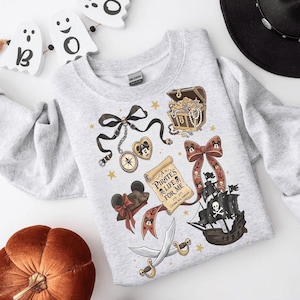 Könnte beinhalten: Ein hellgraues Sweatshirt mit einem Piraten-Design, darunter eine Schatzkiste, ein Piratenschiff und der Text "Pirate's Life For Me". Das Design umfasst einen Kompass, eine Minnie-Mouse-Schleife und gekreuzte Schwerter. Das Sweatshirt ist mit goldenen Sternen verziert.