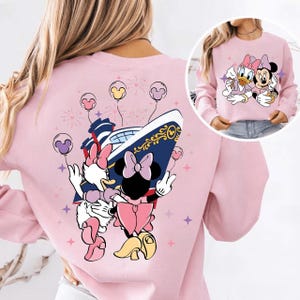May include: A pale pink sweatshirt with a cartoon illustration of Minnie Mouse and Daisy Duck. The back of the sweatshirt features a cruise ship design with Minnie and Daisy, balloons, and fireworks.