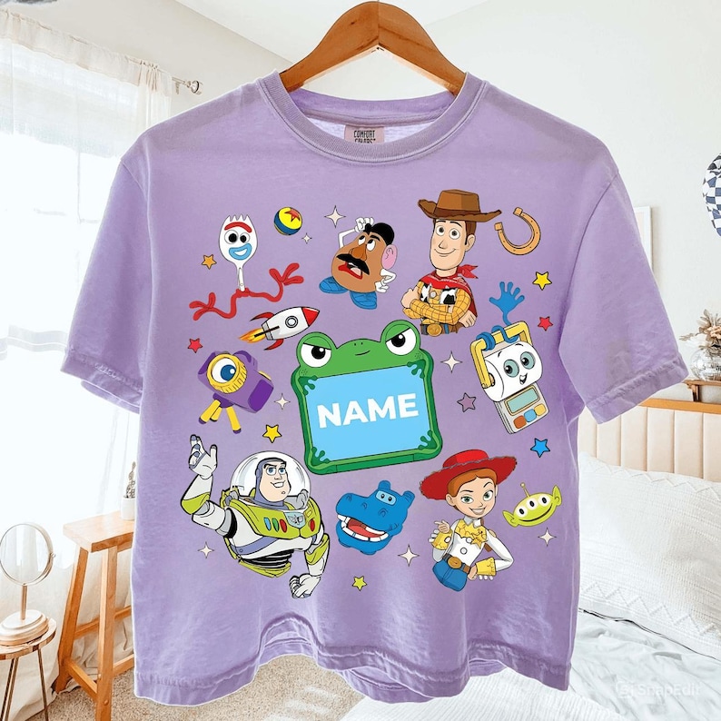 May include: A lilac-colored t-shirt featuring a colorful graphic of characters from the animated movie Toy Story. The design includes Woody, Buzz Lightyear, and other characters, along with the word "NAME" in a green frame. The shirt is hanging on a wooden hanger.