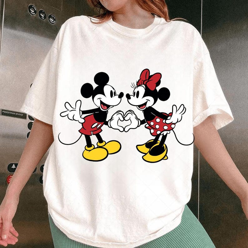 May include: Cream-colored t-shirt featuring a graphic of Mickey Mouse and Minnie Mouse forming a heart with their hands. Mickey wears red shorts and yellow shoes, while Minnie wears a red polka-dot skirt and a red bow.