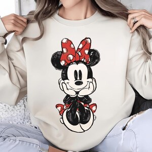 Op de afbeelding: Cr&egrave;mekleurige sweatshirt met een Minnie Mouse-afbeelding. Minnie wordt afgebeeld in een zittende houding, met een rode strik met witte stippen, een zwarte jurk en rode schoenen. Het ontwerp heeft een licht verweerde, vintage uitstraling.