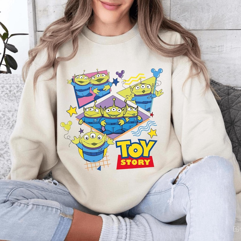 May include: Cream-colored sweatshirt featuring a graphic of the Toy Story aliens. The design includes the words "TOY STORY" in red and yellow, with the aliens in blue uniforms and green skin. The design also includes stars and Mickey Mouse silhouettes.