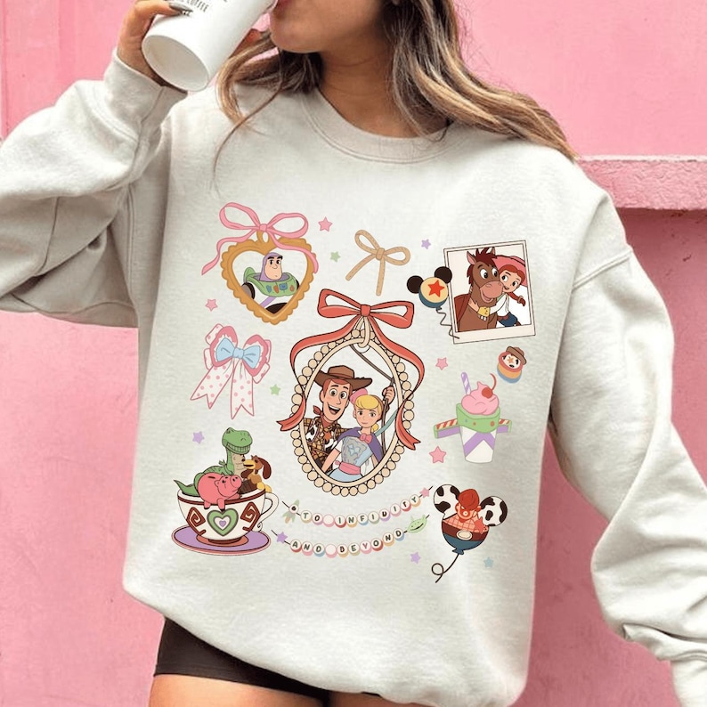 May include: A light gray sweatshirt featuring colorful cartoon graphics. The design includes images of characters from a popular animated movie, such as Woody, Buzz Lightyear, and others, along with decorative bows and stars. The phrase "to infinity and beyond" is also included.