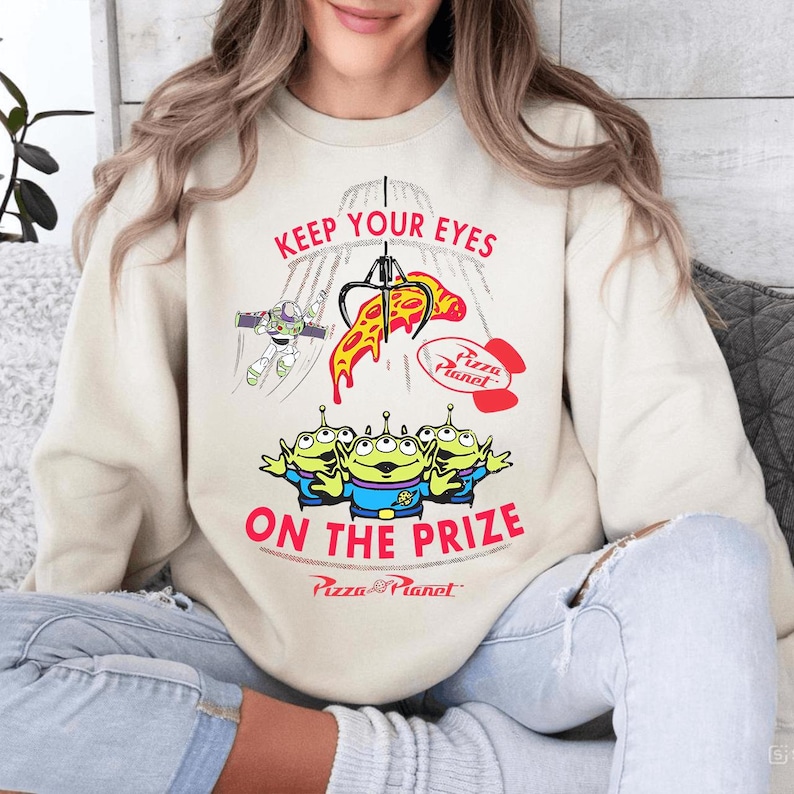 May include: Cream-colored sweatshirt featuring a graphic from the movie Toy Story. The design includes the text "KEEP YOUR EYES ON THE PRIZE" with images of the Pizza Planet claw machine, Buzz Lightyear, and the Little Green Men.