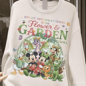 May include: Cream-colored sweatshirt featuring a colorful graphic for the Epcot International Flower & Garden Festival 2026. The design includes Disney characters like Mickey, Minnie, Donald, and Goofy, along with a dragon and other floral elements.