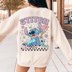 May include: Cream-colored sweatshirt featuring a graphic of the character Stitch with the word "Stitch" in a retro font. The design includes Stitch, flowers, and other cartoon elements. The sweatshirt has a crew neck.