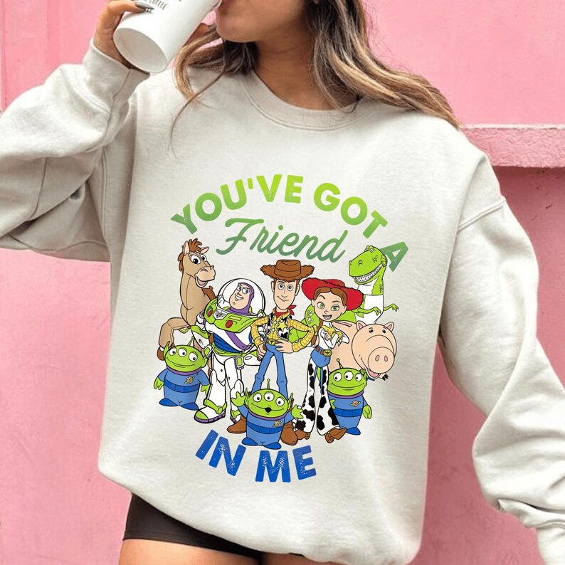May include: A light grey sweatshirt featuring a colorful graphic of characters from the animated film "Toy Story." The design includes Woody, Buzz Lightyear, and other characters, with the text "YOU'VE GOT A FRIEND IN ME" in green and blue.
